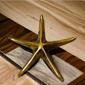 May include: A gold-toned metal starfish sculpture with a textured surface. The starfish is sitting on a wooden surface.