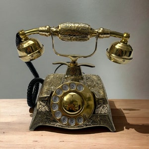 May include: A gold rotary phone with an ornate design and a black cord. The phone has a round dial with numbers 1 through 0.