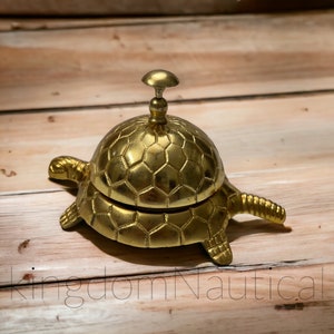 May include: A gold-colored turtle-shaped service bell with a hexagonal pattern on the top and bottom. The bell is on a wooden surface.