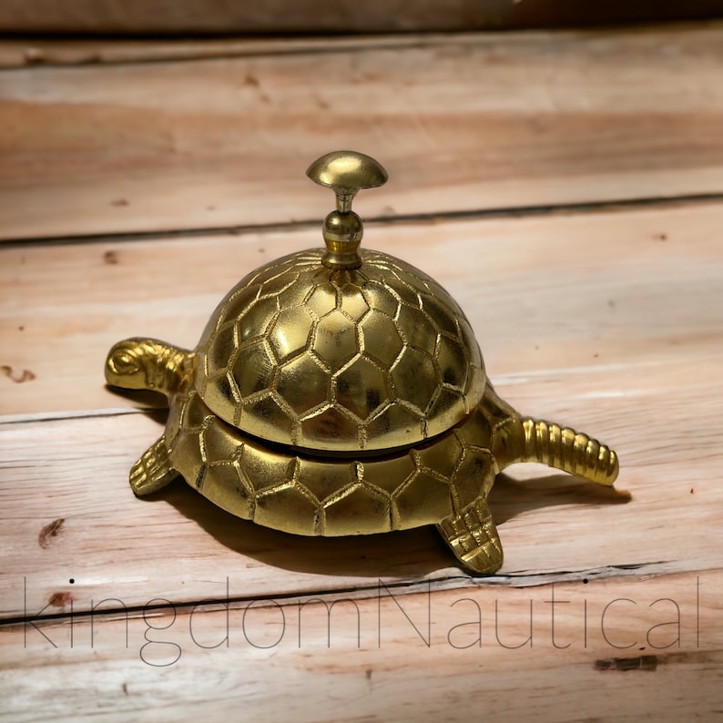 May include: A gold-colored turtle-shaped service bell with a hexagonal pattern on the top and bottom. The bell is on a wooden surface.
