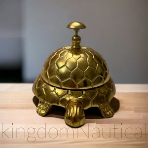 May include: A gold-colored turtle-shaped service bell with a hexagonal pattern on the top. The bell is made of metal and has a shiny finish.