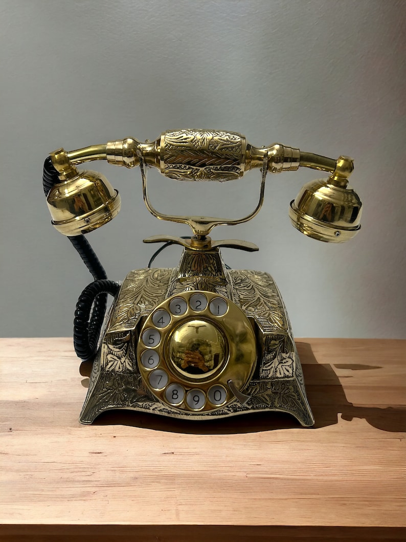 May include: A gold rotary phone with an ornate design and a black cord. The phone has a round dial with numbers 1 through 0.
