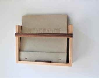 May include: A wall-mounted record holder crafted from light-coloured wood, featuring a dark brown accent. A record album is partially visible, with the text "JOUGH DAWN BAKER / VADE". The holder is fixed to a white wall.