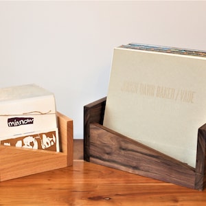 May include: Two wooden record storage boxes. The box on the left is made of light wood and has the word "minnow" printed on the front. The box on the right is made of dark wood and has the text "JOUJOU DAWN BAKER / VADE" printed on the front.