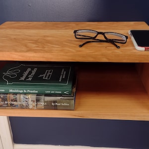 May include: A wooden wall shelf with a small open shelf. The shelf is holding two books, a pair of glasses, and a cell phone. The books are "Moldings in Practice" and "Essential Woodworking Tools".