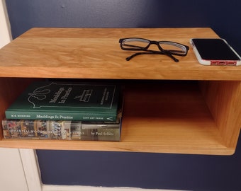 May include: A wooden wall shelf with a small open shelf. The shelf is holding two books, a pair of glasses, and a mobile phone. The books are "Moldings in Practice" and "Essential Woodworking Tools".