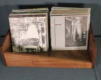 May include: A wooden record stand with a sloping shelf, holding a stack of vinyl records. The record covers feature black and white images and the text "Eat Off The Line" and "Jawbreaker Chesterfield King".