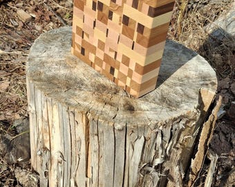 May include: A rectangular end-grain chopping board, featuring a mosaic pattern of light and dark brown wood blocks. The board is displayed on a weathered tree stump.