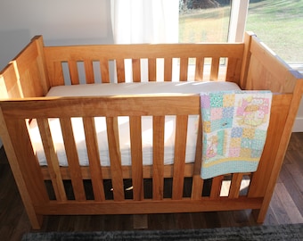 May include: A wooden cot with a white mattress and a colourful patchwork quilt. The cot is made of natural wood and has a simple design.