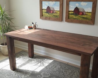 May include: A rectangular, dark brown wooden table with four sturdy legs. The table is in a well-lit room with a window, a potted plant, and two framed paintings of red barns. The table is approximately 152 cm long.