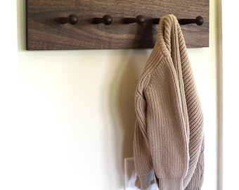 May include: A rectangular dark brown wooden wall-mounted coat rack featuring five evenly spaced pegs. A beige knit sweater hangs from the rightmost peg. The rack is mounted horizontally on a plain wall above a white electrical light switch plate.