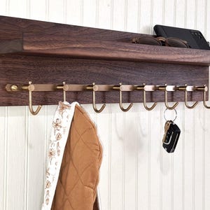 May include: A dark brown wooden wall mounted shelf with a small upper shelf and eight brass hooks. A brown and white patterned scarf hangs from one of the hooks. A set of keys hangs from another hook.