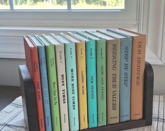 May include: A selection of vintage children's books in a dark wooden book holder. The books feature colourful spines with titles such as "First Steps" and "Busy Times." The books are displayed on a checkered tablecloth near a window.