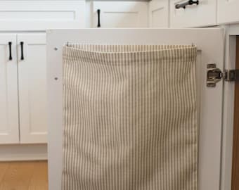 May include: A beige and white striped fabric bag hangs inside a white kitchen cabinet door. The cabinet door has black handles and is open, revealing the interior. The background shows a white kitchen with a wooden shelf and a stainless steel tap.