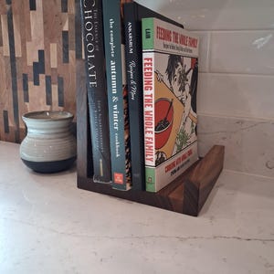 May include: A wooden cookbook holder with four books. The books include "The Essence of Chocolate," "The Complete Autumn & Winter Cookbook," "Recipes & More," and "Feeding the Whole Family." The holder is on a white countertop.