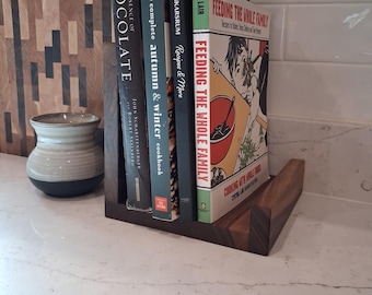 May include: A wooden cookbook holder with four books. The books include "The Essence of Chocolate," "The Complete Autumn & Winter Cookbook," "Recipes & More," and "Feeding the Whole Family." The holder is on a white countertop.