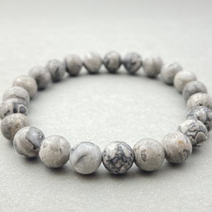 May include: A gray and white patterned stone beaded bracelet. The bracelet is made of round beads with a marbled pattern.