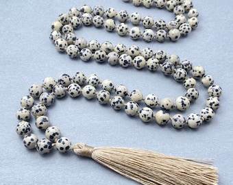 May include: A long, white and black speckled stone mala necklace with a beige tassel.