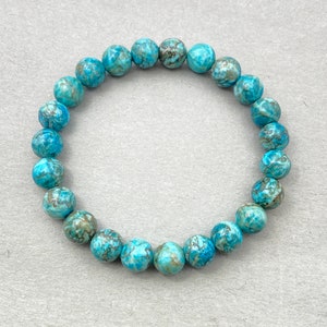 Puede incluir: Un brazalete de piedras preciosas azul turquesa con un patr&oacute;n de m&aacute;rmol. El brazalete est&aacute; hecho de cuentas redondas y est&aacute; enhebrado en un cord&oacute;n el&aacute;stico transparente.