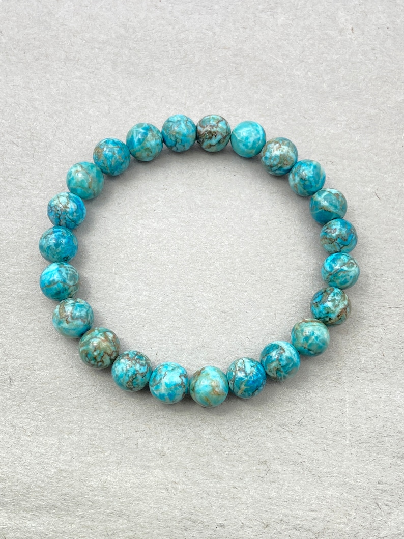 Puede incluir: Un brazalete de piedras preciosas azul turquesa con un patr&oacute;n de m&aacute;rmol. El brazalete est&aacute; hecho de cuentas redondas y est&aacute; enhebrado en un cord&oacute;n el&aacute;stico transparente.