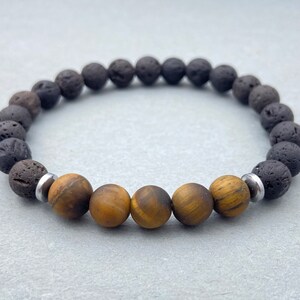 Op de afbeelding: Een armband met donkergrijze lavasteen kralen en goudbruine tijgeroog kralen. De armband is geregen op een elastisch koord en bevat zilverkleurige metalen spacer kralen. De armband is ontworpen voor dagelijks gebruik.