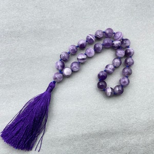 May include: A purple amethyst mala with a purple tassel. The beads are round and have white streaks.