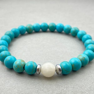 Pode incluir: Um bracelete de contas turquesa e brancas com detalhes prateados. O bracelete é feito com contas redondas e tem uma única conta branca no centro.