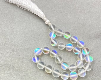 Op de afbeelding: Een witte kralenketting met een witte kwast. De kralen zijn iriserend en hebben een regenboogkleurige glans.