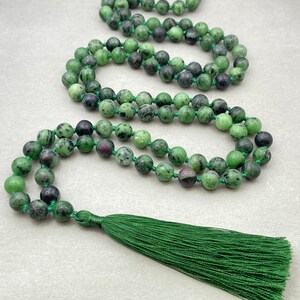 Könnte beinhalten: Eine grüne und schwarze Perlenkette mit einem grünen Quastenanhänger. Die runden Perlen scheinen aus Stein zu sein. Die Kette ist auf grünem Faden aufgefädelt. Die Quaste besteht aus grünem Faden.