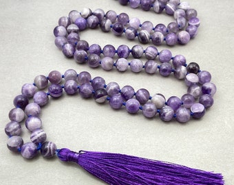 May include: A long necklace made of purple and white striped beads, strung on blue thread. The necklace ends with a deep purple tassel. The beads are round and appear to be made of stone or a similar material.