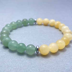 Może przedstawiać: Bransoletka z koralików z gradientem kolorów. Posiada jasnozielone koraliki, które przechodzą w żółte. Okrągłe koraliki wydają się być wykonane z gładkiego, polerowanego materiału. Bransoletka jest nawleczona na elastyczną gumkę ze srebrnymi przekładkami.