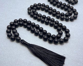 May include: A long strand of matte black spherical beads arranged in a double loop, featuring a matching black thread tassel at the end. The beads are uniform in size and texture, creating a continuous circular pattern connected by a dark cord.