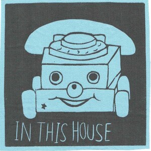 May include: A square, light blue and black fabric patch with a cartoon telephone design. The telephone has a smiling face and the text "IN THIS HOUSE" in white letters.