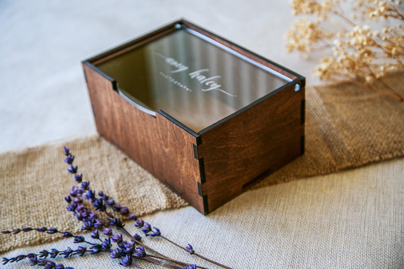 May include: A rectangular wooden box with a dark brown finish and a clear acrylic lid. The lid features the white printed text "amy haley PHOTOGRAPHY" in a script and sans-serif font. The box has visible interlocking joints at the corners.