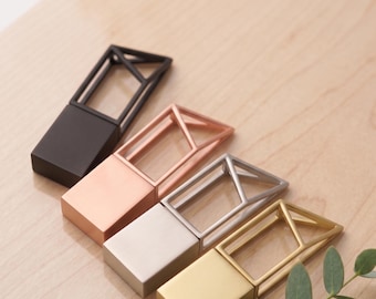 May include: Four geometric metal bottle stoppers in black, copper, silver, and gold, arranged diagonally on a light wood surface. Each stopper features a rectangular base and a geometric cage design. A sprig of green leaves is in the lower right corner.