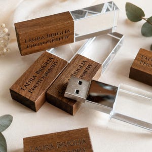 Puede incluir: Varias memorias USB rectangulares con una tapa de madera marrón oscuro grabada con el texto LAURA BRIGLIA PHOTOGRAPHY y un cuerpo de acrílico transparente. Una unidad está abierta, revelando un conector USB de metal plateado.
