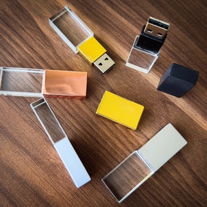 May include: A collection of six crystal USB flash drives in various colors, including gold, silver, and black. The drives are arranged on a wooden surface.