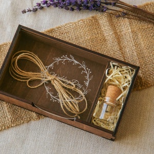 Op de afbeelding: Een houten doos met een doorzichtig deksel, met daarin een klein flesje met een kurkstop en een stuk touw dat in een strik is geknoopt. De doos is gegraveerd met de woorden "Wedding Day" en een krans van bloemen.