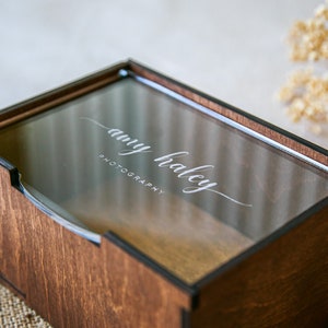 May include: A rectangular wooden box with a dark brown finish and a clear acrylic sliding lid. The lid features the white engraved text "amy haley PHOTOGRAPHY" in a combination of script and sans-serif fonts. The box has visible joinery.