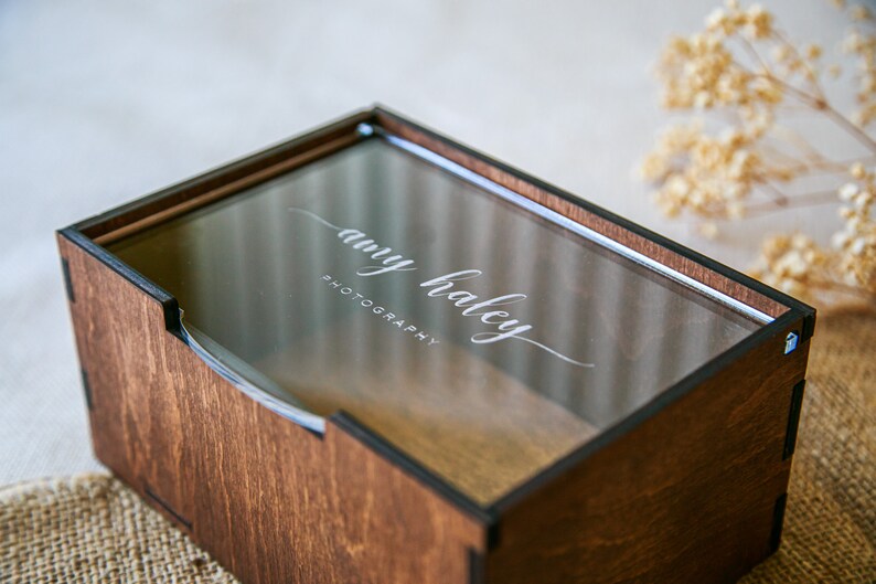 May include: A rectangular wooden box with a dark brown finish and a clear acrylic sliding lid. The lid features the white engraved text "amy haley PHOTOGRAPHY" in a combination of script and sans-serif fonts. The box has visible joinery.