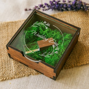Puede incluir: Una memoria USB de madera dentro de una caja cuadrada de madera con tapa de cristal transparente. La caja está llena de musgo verde y tiene las palabras "Wedding Day" grabadas en el cristal. Una ramita de lavanda está en el fondo.