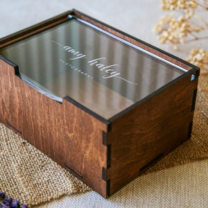 May include: A rectangular dark brown wooden box with a clear acrylic sliding lid. The lid features the white engraved text "amy haley PHOTOGRAPHY" in a script and sans-serif font. The box has visible interlocking joints at the corners.