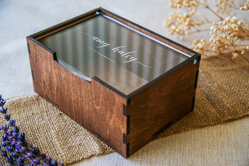 May include: A rectangular dark brown wooden box with a clear acrylic sliding lid. The lid features the white engraved text "amy haley PHOTOGRAPHY" in a script and sans-serif font. The box has visible interlocking joints at the corners.