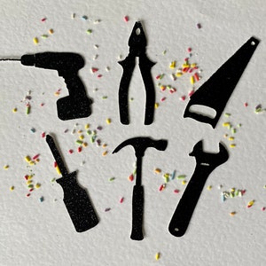 May include: A set of six black silhouette tool shapes, including a drill, screwdriver, wrench, hammer, saw, and pliers, on a white background with colourful sprinkles.