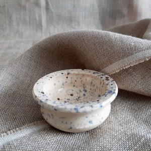 May include: A small white ceramic bowl with blue and brown speckles. The bowl is round and has a slightly flared rim.