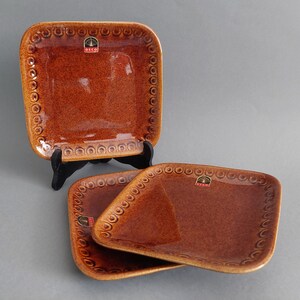 Può includere: Tre piatti quadrati in ceramica marrone con un bordo decorativo e un piccolo logo che dice "DECO" al centro.