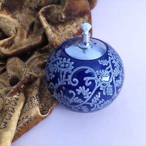 Op de afbeelding: Een blauwe keramische olielamp met een zilveren deksel en een witte lont. De lamp is versierd met een wit bloemenpatroon.