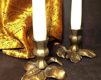 May include: Two brass candle holders with leaf-shaped bases and white candles. The candle holders are on a dark brown surface.