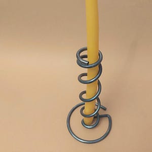 Op de afbeelding: Een metalen kandelaar met een spiraalvormig ontwerp. De houder is gemaakt van donkergrijs metaal en heeft een gele kaars. De kandelaar is een uniek stukje woondecoratie.