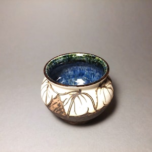 Puede incluir: Un pequeño cuenco de cerámica blanca con un borde marrón y un glaseado azul y verde en el interior. El cuenco tiene un diseño tallado de hojas en el exterior.
